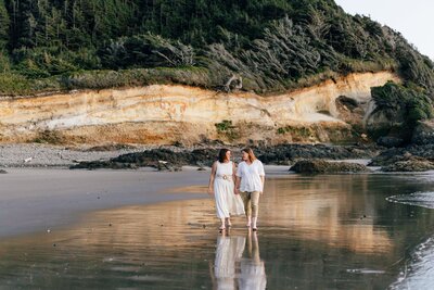 Two brides wearing white walk barefoot while holding hands as the water casts a reflection on the sand during their Oregon Coast adventure elopement. | Erica Swantek Photography
