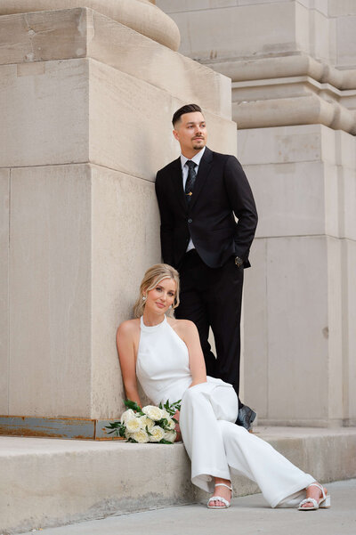 an editorial style photo of a bride in a pantsuit sitting holding her groom's hand outside the Chateau Laurier taken by Ottawa wedding photographer JEMMAN Photography