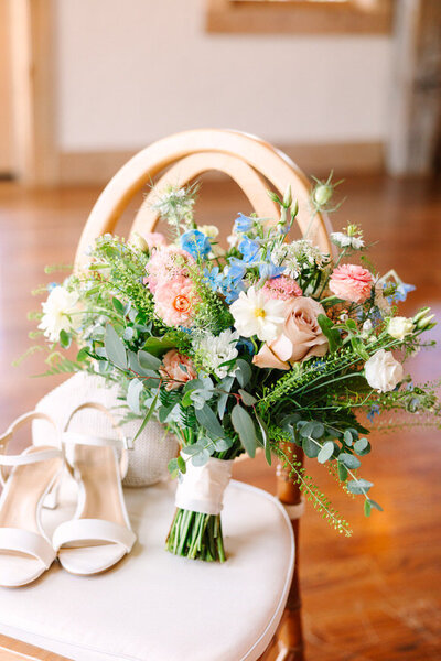 Bridal bouquet with roses and greenery alongside white wedding shoes on a chair, captured by a North Carolina wedding photographer.