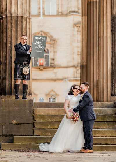 A newlywed couple sharing a kiss on stone steps in front of a historic building, with guests watching nearby, beautifully captured by a UK wedding photographer and videographer.