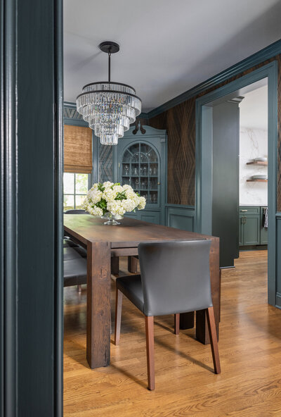 crystal and dark metal chandelier over dark wood rough hewn rectangular table with gray leather dining chairs, oak wood floor with grayish green painted chair rail and trim paint color, traditional built-in hutch, greenish gray background with large gold diamond pattern grasscloth wallpaper