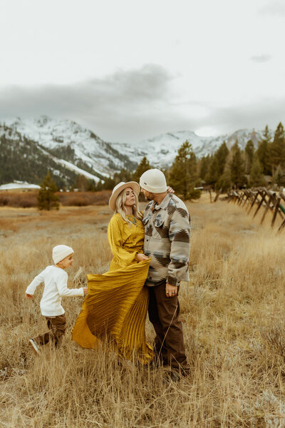Mom and dad are snuggled together standing up as their boy runs around them in a field with snowy mountains behind them