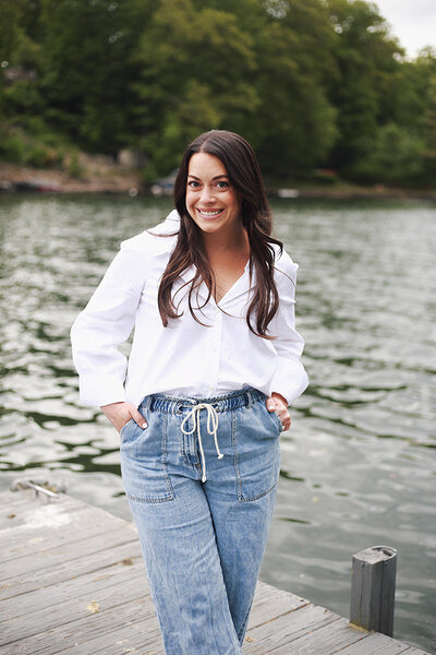 Emily Austin in jeans stands on a dock, looking out over the water, with a serene expression on her face.