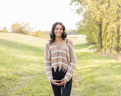 high school female senior, with long black hair, standing in grass with a tree line next to her, looking at the camera smiling, wearing black leggings, and light tan and white stripped shirt, fingers interlocked in front of her
