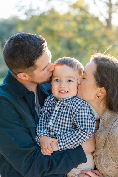 Parents kiss their toddler on each cheek while he smiles, photographed outdoors by a Worcester family photographer.