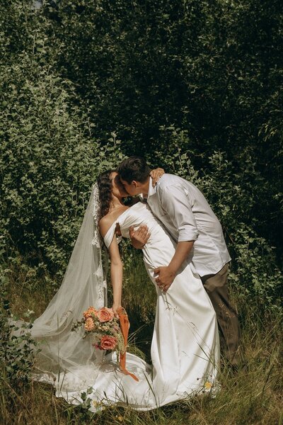 Couple sharing a kiss in a sunlit meadow with bride holding bouquet