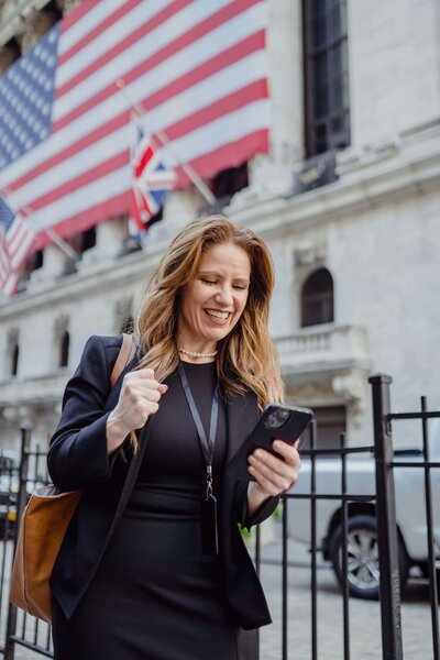 A professional woman in a black outfit, smiling and holding her phone triumphantly with a fist pump, standing outside a building adorned with large American flags.