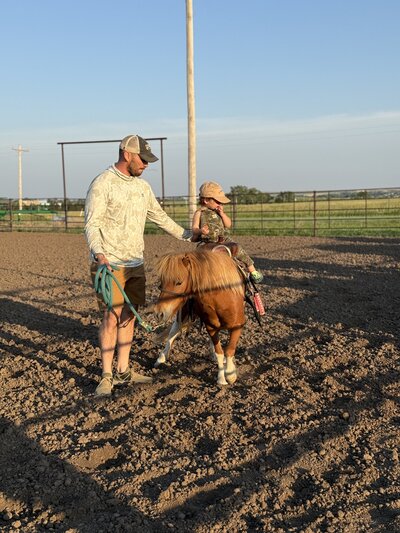 father and child leading a pony across a muddy pen at golden hour on the farm

