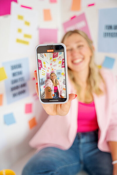 Kat Murphy in a pink suit sitting with her arm on a disco ball holding her phone in front of a pink background
