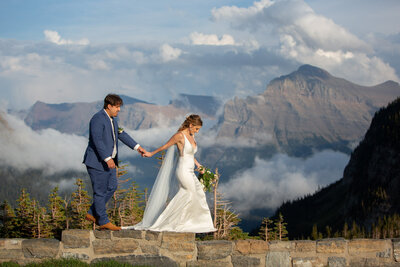 Bride and groom walk hand in hand on a mountain overlook, surrounded by dramatic peaks and swirling clouds.