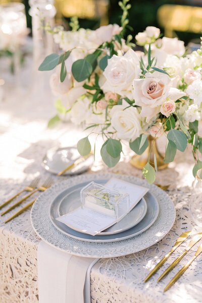Wedding reception table setting, 3 white plates stacked together, soft pink roses, lacy table cloth, and gold cutlery.