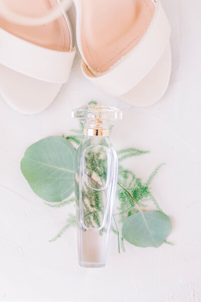 Aerial view of clear and gold perfume bottle laying on top of eucalyptus greenery in front of nude high heels taken by New Orleans wedding photographer Elizabeth Collins