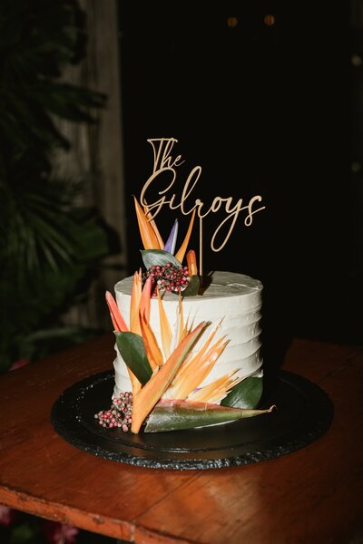 A white, single-tier wedding cake adorned with tropical flowers, including bird of paradise, and a custom cake topper reading ‘The Gilroys.’ The cake sits on a dark table with a tropical backdrop. Keywords: destination wedding planner, tropical wedding decor, luxury destination weddings.