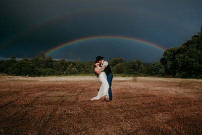 Bride and groom sharing a kiss in an open field under a double rainbow, with dramatic storm clouds in the background during their elopement