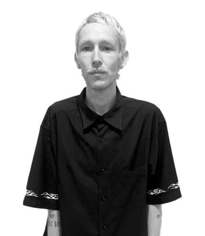 Male Art Director with short platinum hair, tattoos, and a black button-up shirt, embodying creative and edgy hair artistry.