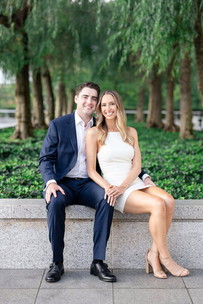 A couple is sitting on a stone ledge outside, surrounded by greenery and trees. The man wears a navy suit, and the woman is in a white dress. Both are smiling at the camera.