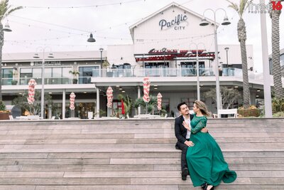 Bride to be sits on her fiance's lap as he sits on the steps to Pacific City in Huntington Beach