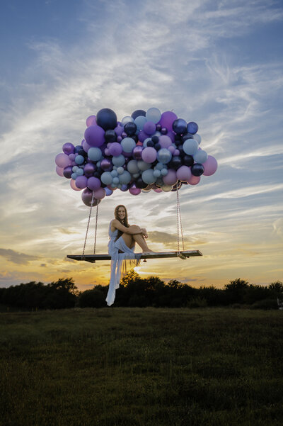 A senior girl poses on a lifted bench with a purple and blue balloon arch at sunset.