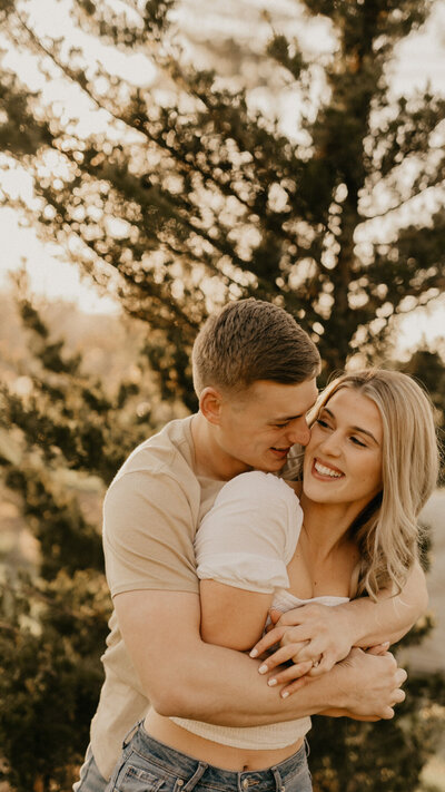 Young man wraps up his fiance in a huge embrace from behind as he looks over her shoulder and smiles at her. His stunning blonde haired fiance looks back at hime with a big smile while grabbing his hands.