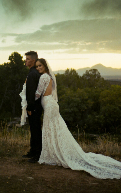 A 35mm film image of a couple on their wedding day facing the camera with their arms around each other. There is a mountain in the back and it's sunset.