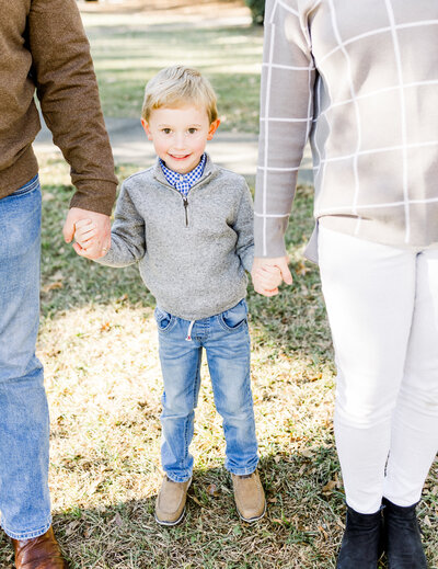 A young boy with blonde hair and a bright smile stands between his parents, holding their hands during an outdoor family session. Dressed in a cozy sweater and jeans, he radiates warmth and happiness, capturing the natural and timeless style of a Birmingham family photographer.