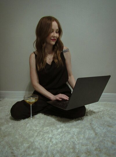A Showit Design Partner with long hair sits cross-legged on a light carpet, using her laptop to explore branding and website design, with a glass of drink placed beside her.