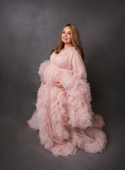 Expectant mom in organza and tulle gown stands for maternity photos in Brooklyn, NY photo studio. Mom has her hands resting on her bump and smiling at the camera.