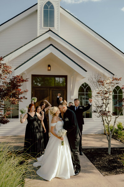 bride and groom kissing while their wedding party cheers in the background in front of white wedding chapel at The Springs