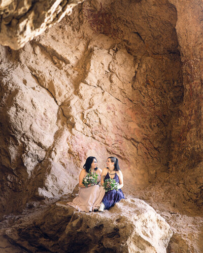 Brides sit together with their bridal bouquets in a cave during their Texas adventure elopement.