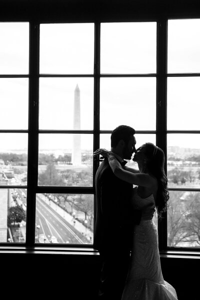 A couple embraces in silhouette against a large window, with the Washington Monument visible in the background. The scene captures a romantic and intimate moment, with a cityscape view beyond.