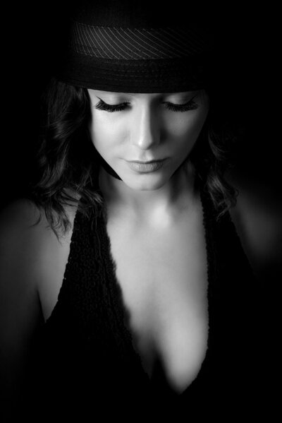 Striking low-key studio portrait of a woman in a black dress and fedora, lit in a dramatic cinematic style.