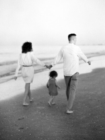 Mother and father hold hands above young daughters head as they walk along the beach