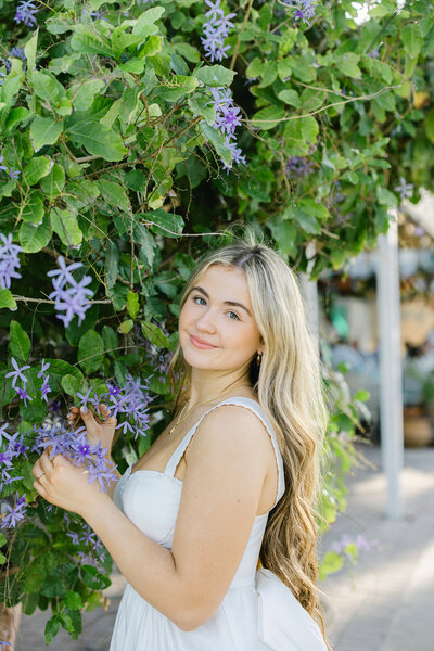 High School Senior Portrait Wearing A White Dress Standing By Purple Flowers