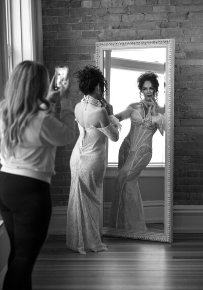 A woman in a lace gown poses confidently in front of a mirror while the hair and makeup artists takes behind the scene shots.