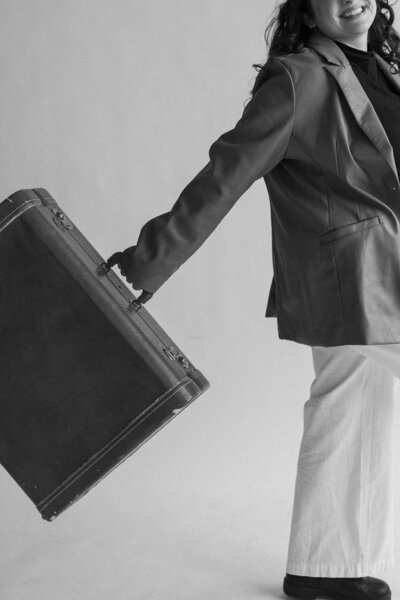 A smiling woman in a stylish blazer and white pants holds a vintage suitcase, poised confidently against a light gray background.