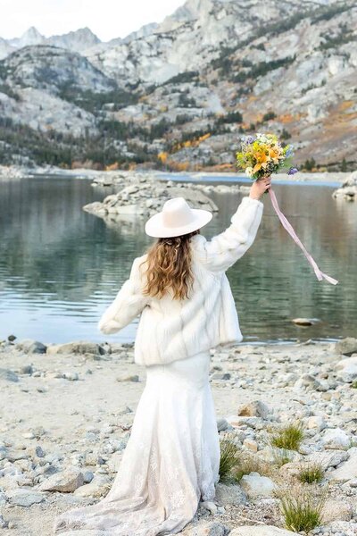 A bride raises her bouquet overhead in celebration in front of a beautiful lake view on her mammoth wedding day.