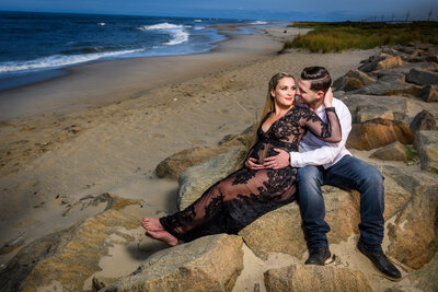an expectant couple seated on large rocks at the beach; the pregnant woman in a lace dress is embraced by her partner, with the ocean waves and sandy shore in the background