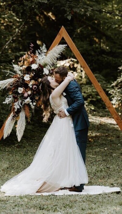 A bride and groom kiss in front of a boho-inspired floral arch, representing wedding flowers designed by Flowers and Thyme.