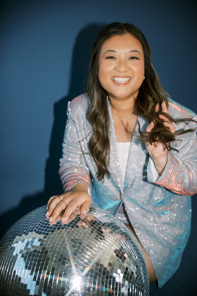 Monique Anne in a sparkly blazer sitting next to a disco ball in front of a blue background