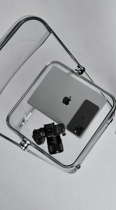 A collection of modern tech devices—a silver MacBook, a black iPhone, and a Sony digital camera—are arranged on the clear seat of a transparent acrylic chair against a clean, light-colored background