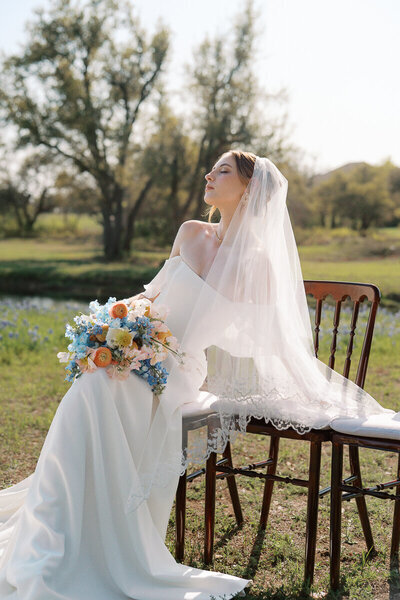 Bridal portrait in the field at Ma Maison wedding venue near Austin, Texas. The bride is seated on a wooden chair in a lace-trimmed veil, holding a pastel spring bouquet, basking in soft sunlight.