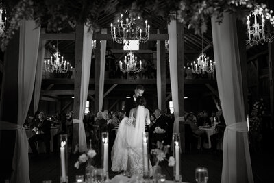 A black and white photo of a bride and groom sharing their first dance in an elegant barn venue surrounded by chandeliers and candlelight.