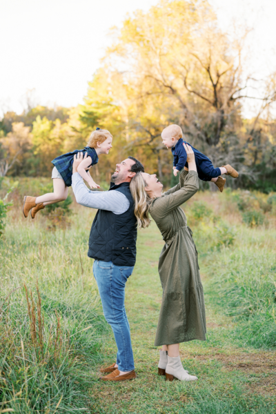 Mom holds up toddler girl and dad holds baby boy during playful Fall family photos