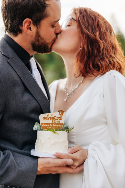 couple is sharing a kiss while holding a small cake that has their names and date on the sign. 