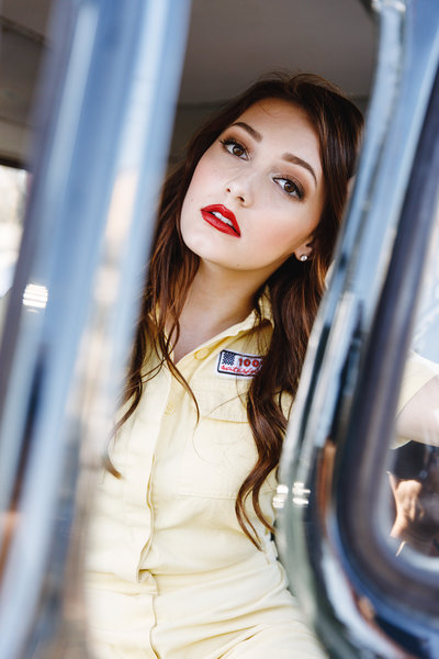  Vintage-inspired senior portrait of a young woman with red lipstick and loose waves, framed creatively through a car window while wearing a retro yellow button-up.

