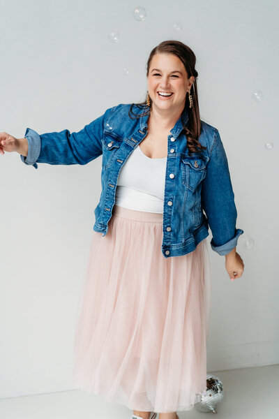 Female in jean jacket and light pink skirt kicking leg up on a couch.