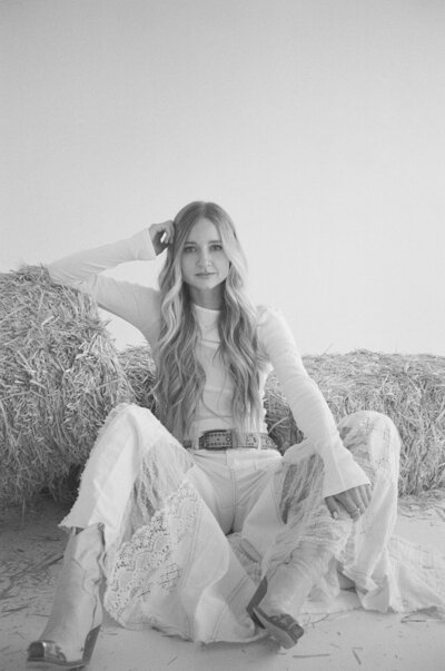 woman wearing all white sitting on the floor and leaning against a hay bale