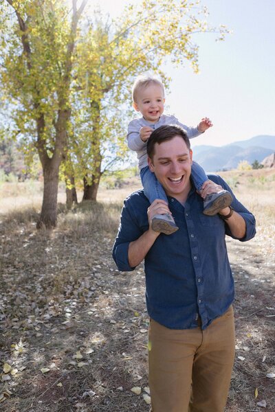 A toddler rides on his dad's shoulders in front of a yellow sunlit tree.