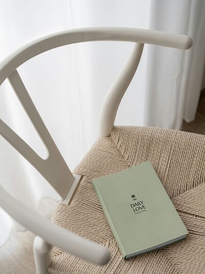 MPR Ministries branded devotional book titled “Daily Love” in soft sage green with the logo icon on the cover, placed on a neutral woven chair to highlight the peaceful, faith-centered tone of the brand.