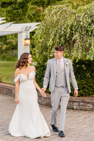Bride and groom walking in front of a beautiful white pergola holding hands looking at each other.